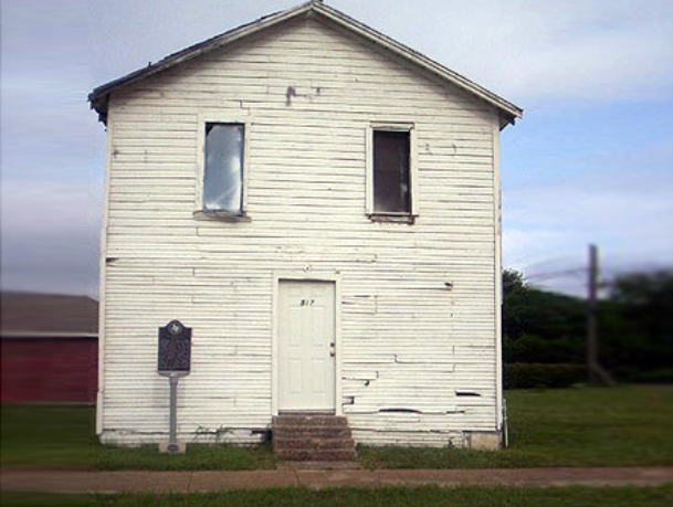 A gable front two story white clapboard building fills the frame. There are several steps to a white door and two rectangular windows on the second story. A THC Historic Marker is in front.